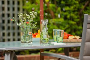 View of the outside table with a jar with flowers, a carafe with water and cucumber and two glasses. A bowl with clementines and lemons and a cut apple on the right