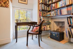 Image of a library corner with a desk facing a window with a chair and the walls to the right filled with books