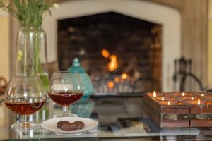 Image of a lit fireplace in the background and a coffee table with candles, a dish with chocolate bonbons and a couple of glasses with hot brandy