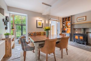 Image of a dining room with a table and chairs. There is a shelving unit to the right and two windows.