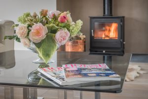 Image of a coffee table with magazines, a vase with flowers and a lit fireplace in the background